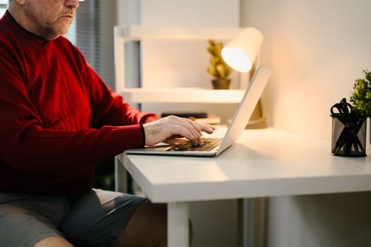 Close up of elderly man using laptop at minimalist desk under desk lamp.