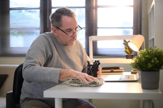 Senior man cleaning his desk at home office, wiping surface with cloth.