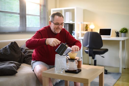 Mature male enjoying a slow coffee brewing process with a kettle and dripper in living room.
