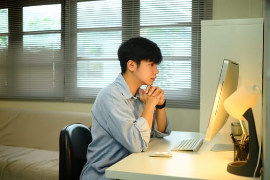Young man working from home, sitting at desk and concentrating on desktop computer screen.