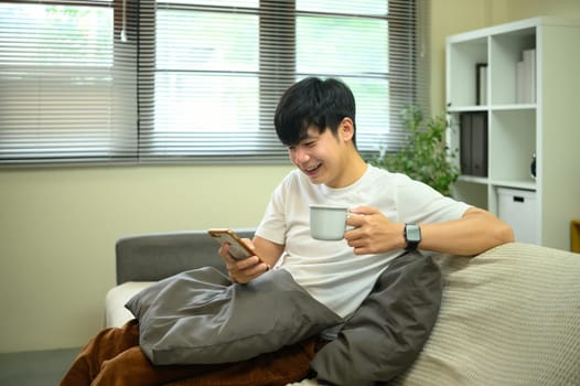 Asian male smiling while checking smartphone and holding a coffee mug on sofa.