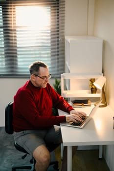 Senior man in red sweater working on a laptop at minimalist desk under desk lamp.