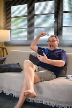 Excited senior man celebrating online success while holding smartphone and credit card.