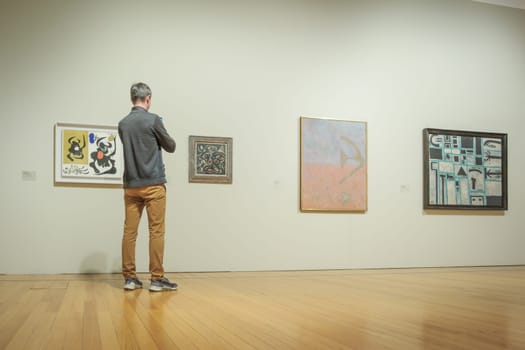 Jan 13, 2024 - Lisbon, Portugal: visitor in gray jacket and tan pants observe abstract artworks displayed on white walls of Berardo Collection Museum, deeply engaged in contemplation in artistic details. Wooden floor, serene atmosphere, minimalistic