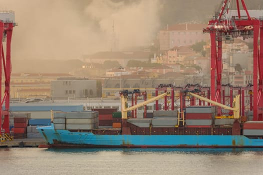 Blue container ship docked at Lisbon port, red cranes and stacks of shipping containers on deck, city buildings and church spire in evening haze and fire smoke in background. Risk of fires in summer, drought and heatwave, danger, damage, Portugal