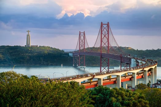 Scenic view of Lisbon iconic 25 de Abril Bridge over Tagus River with Cristo Rei or Christ the King statue in background at sunset. City traffic, lush greenery, tranquil river, famous landmarks, red suspension bridge, colorful sky, travel destination