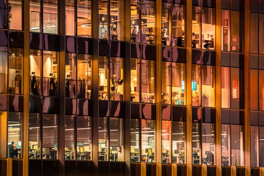 Contemporary office building at night with large glass windows revealing warmly lit workspaces. Employees are visible at desks and computers, active business atmosphere and modern corporate environment, after sunset, modern work environment
