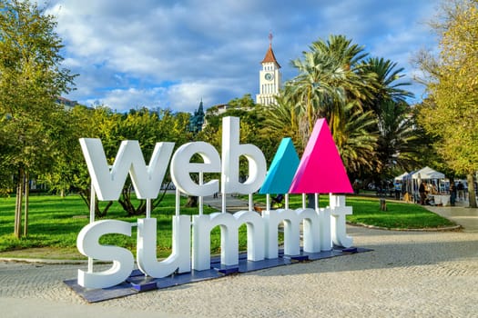 Nov 13, 2024 - Lisbon, Portugal: Large sign displaying Web Summit logo in lively park filled with greenery, historic tower in background. Major annual technology conference internet technology, emerging technology, marketing, AI solution, networking