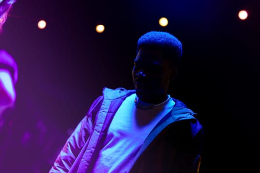 A young man stands in a vibrant atmosphere illuminated by colorful lights, showcasing a modern jacket. The energy hints at a lively event, possibly a concert or gathering.