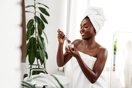 A woman with a towel on her head smiles as she applies skincare in a bright and modern bathroom. Fresh plants enhance the serene atmosphere, creating an inviting space for self-care.