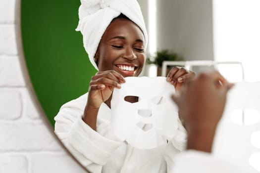 A woman with a towel on her head is happily holding a facial mask in a bright bathroom. The scene captures her excitement to indulge in self-care and beauty routine.