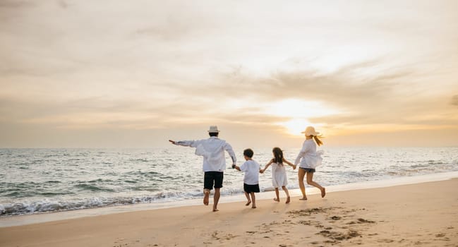 A happy Asian family seen from behind enjoys sunny day at beach. Parents and daughters bond running jumping and pretending to fly with arms outstretched. carefree family moment by sea.