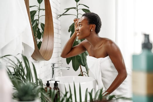 A woman applies skincare products in a stylish bathroom filled with greenery. The sunlight highlights her careful routine while she enjoys a moment of self-care.