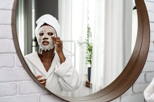 In a well-lit bathroom, a woman in a bathrobe applies a facial mask while looking at her reflection in a round mirror. Natural light creates a calming atmosphere.