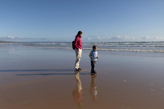 Mother and son enjoying a leisurely walk on the wet sand of a beach at low tide, with their reflections visible in the shallow water