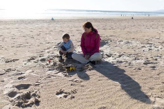 Mother and son enjoying quality time together, sitting on a sandy beach and playing with toys