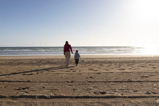 Mother and son enjoying a leisurely walk on a beautiful sandy beach towards the ocean as the sun sets