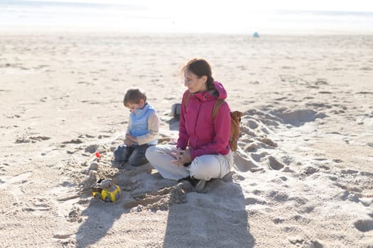 Mother and son enjoying leisure time on sandy beach, building sandcastles and playing with toys