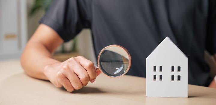 Magnifying glass in hand focuses on a cottage house, symbolizing inspection and real estate appraisal. Ideal for property evaluation, home search, or dwelling assessment in the housing market.