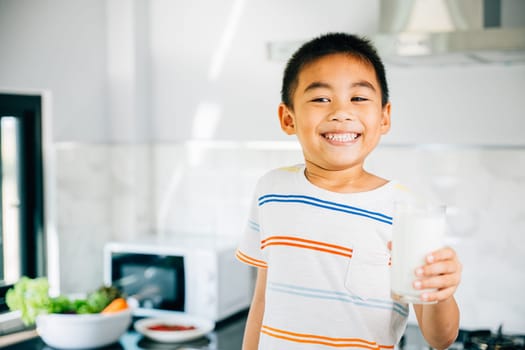 Portrait of adorable Asian preschooler, boy holds milk in kitchen. Smiling son enjoys drink, radiating joy. Happy child savoring calcium-rich liquid, feeling cheerful at home give me.