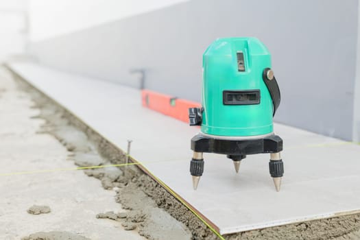 Professional construction tools, green laser level positioned on a tripod for accurate ceramic tile placement on a white and gray floor.
