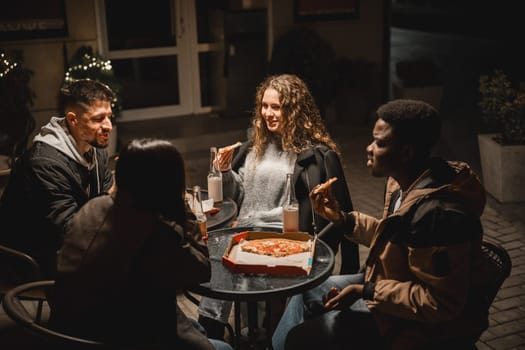 A group of friends share laughter and slices of pizza around a table at night, illuminated by soft lights. Their joyful conversation adds to the cozy atmosphere.