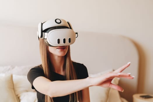 A young woman wearing a virtual reality headset immerses herself in a digital world. She extends her hand as she interacts with the virtual environment in a comfortable setting.