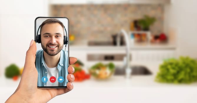 A person holds a smartphone showing a smiling individual on a video call. The background features a cozy kitchen with fresh vegetables, highlighting a casual conversation atmosphere.