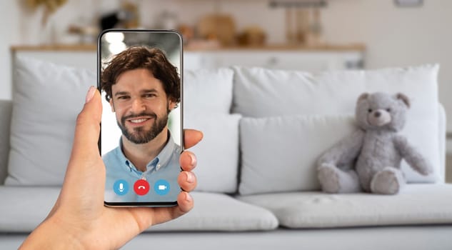 A person participates in a video call, holding a smartphone with a smiling face on the screen. The setting is a cozy living room with a couch and a teddy bear, creating a warm atmosphere.