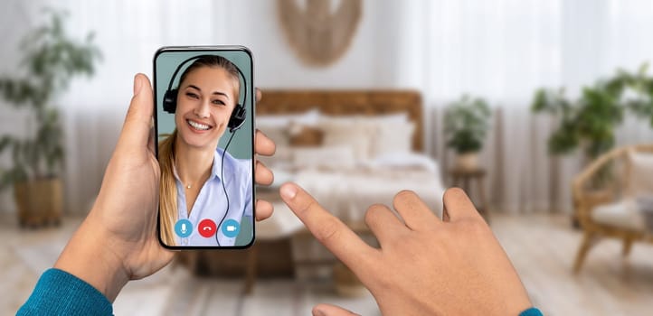 A person is using a smartphone to participate in a video call, interacting with a smiling individual wearing a headset. The setting is a cozy home environment with soft furnishings.