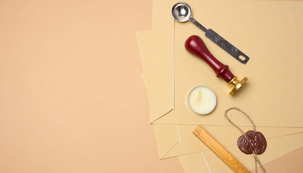 Wax seal kit including wax sticks, spoon, candles, stamp and brown envelopes placed on beige background. Copy space
