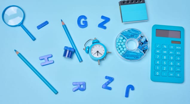 Flat lay of blue stationery, learning items letters, magnifying glass, supplies, and an alarm clock on a blue background. Education, time, and study concept