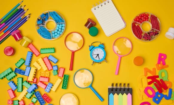 Vibrant collection of colorful school supplies, building bricks, letters, and learning items scattered on a yellow background. Play and education concept.
