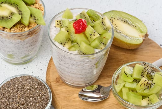 A glass of chia seed pudding layered with kiwi and granola, topped with kiwi slices, sits on a wooden board with ingredients around it