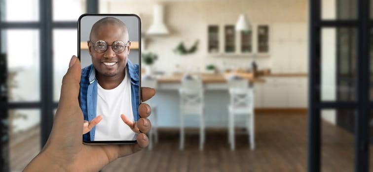 A person holds a smartphone displaying a smiling individual during a video call. The setting features a bright kitchen with wooden floors and a modern, inviting atmosphere.