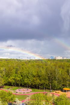 gray rainy clouds and two rainbow over sunlit playground near city park after rain on sunny spring day