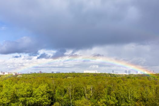 gray rainy clouds and rainbow in blue sky over green city park after rain on sunny spring day