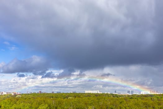 grey rainy clouds and rainbow over green city park after rain on sunny spring day