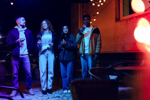 Friends are gathered outdoors at night, holding drinks and enjoying each other's company while surrounded by string lights that create a warm ambiance.