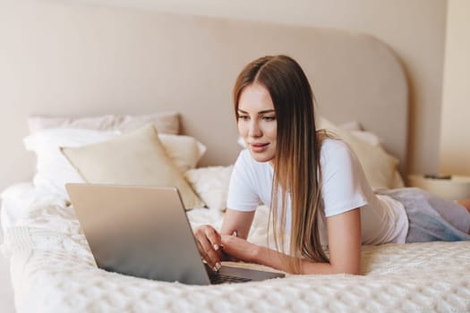 A young woman relaxes on her stomach on a bed, focused on her laptop. Soft natural light fills the modern bedroom, creating a calm atmosphere for productivity.