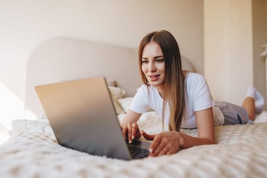 A woman with long hair is comfortably lying on her stomach in bed, engaging with her laptop. Natural light fills the inviting bedroom space, creating a relaxing atmosphere.