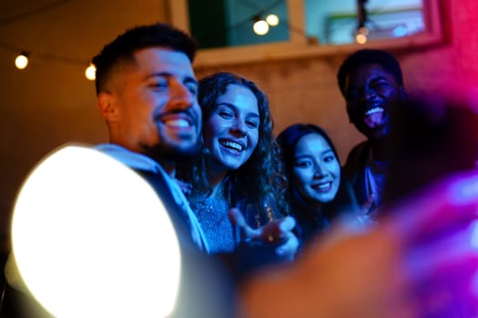 Group of four friends smiles joyfully during a lively nighttime outdoor gathering, surrounded by colorful lights and drinks in a cozy atmosphere.