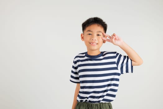 Asian young kid boy showcases optimism and peace by gesturing victory and peace signs in a studio shot with a white background. primary child joyful smile speaks of success, V sign finger