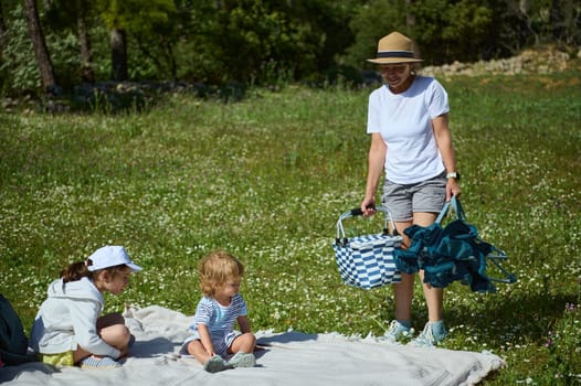 A serene family outing in nature, depicting children seated on a blanket as their mother prepares the picnic, surrounded by greenery and sunshine, embodying outdoor harmony.