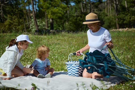 A family with children picnicking on a sunny day in a field surrounded by lush green trees, enjoying quality time in a natural setting.
