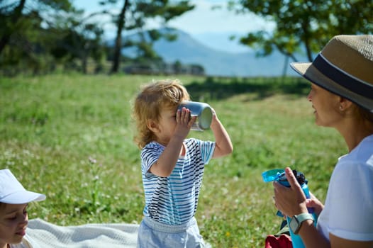 A family picnic on a sunny day amidst beautiful mountainous scenery. A child drinks from a cup while the mother interacts lovingly, embodying the joy of outdoor moments.