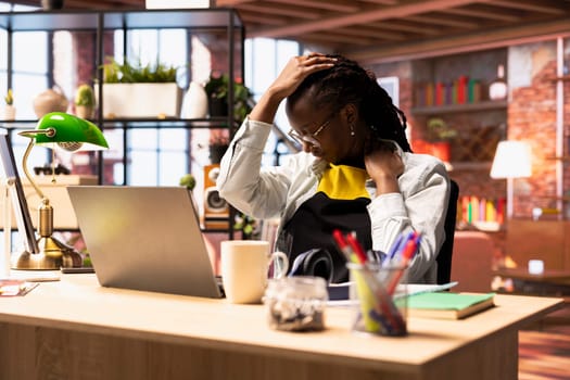 Fatigued woman in home office stretching sore neck after sitting at desk too long, taking break. Exhausted freelancer in apartment doing muscle exercises to relieve painful stinging muscle spasm