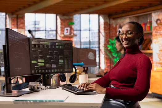 Portrait of upbeat woman working in innovative startup office using programming languages to build application. Jolly African american software development agency IT expert optimizing code on computer