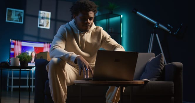 Ambitious black man learning a new language alone in his cozy living room, browsing internet for study platforms and explanations. Adult using language learning software online on laptop. Camera B.