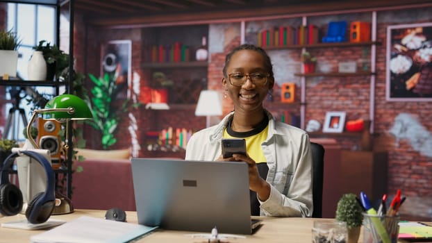 Portrait of jolly woman sitting on chair at home and texting friends on smartphone. Cheerful african american person in living room enjoying leisure time, chatting with mates, camera B